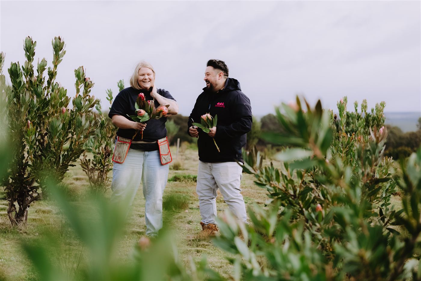 Two of us laughing in the flower paddock, each holding freshly cut proteas